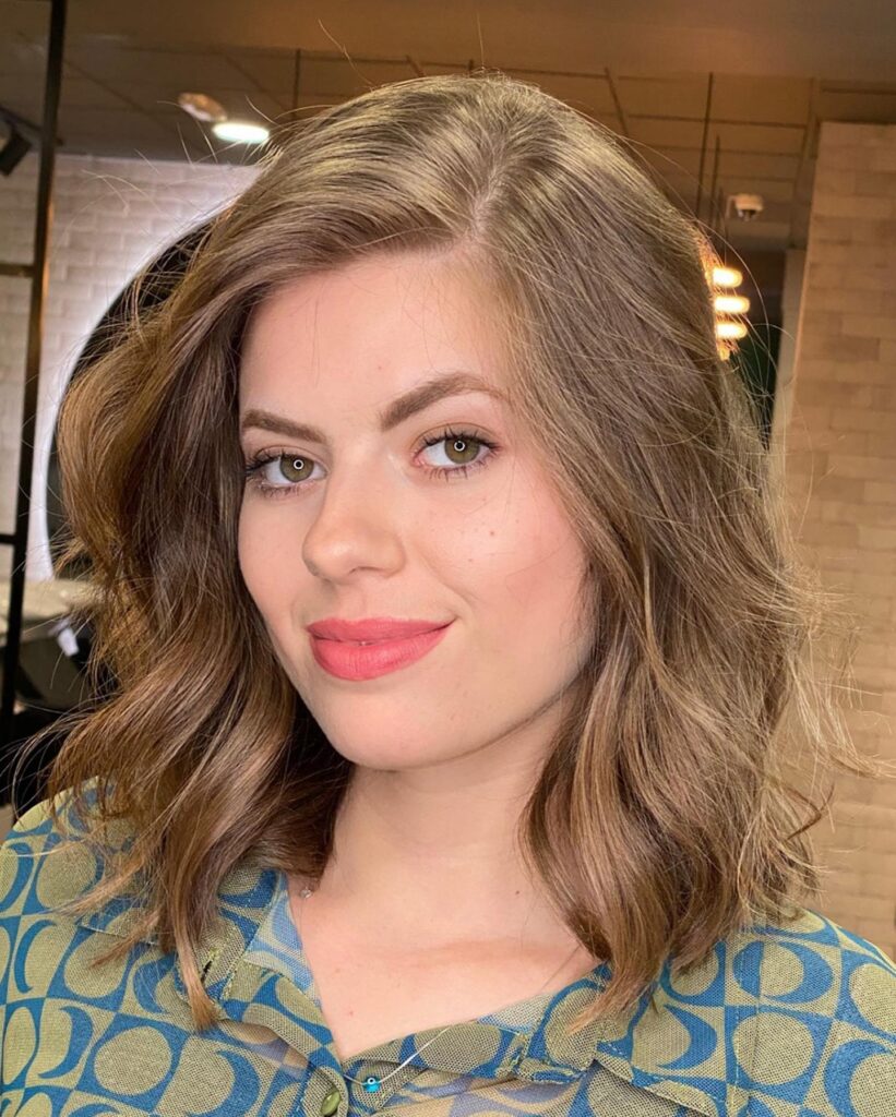 A woman with a voluminous golden brown lob featuring a deep side part and soft tousled waves.