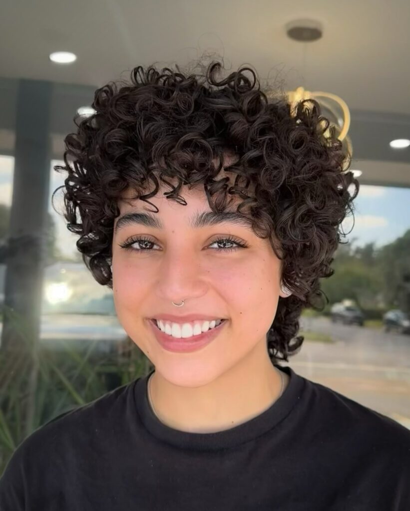 A woman with a voluminous dark curly pixie cut and curly bangs smiling at the camera.