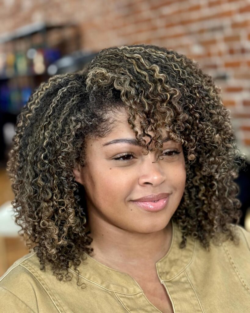 A woman with shoulder-length curly hair featuring blonde highlights and a side-swept curly fringe.