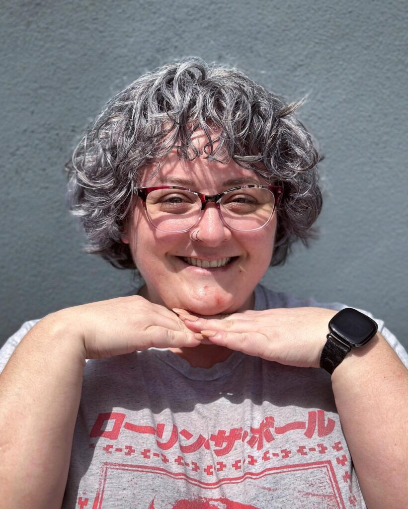 A smiling woman with a short curly silver and gray bob featuring light, wispy bangs.