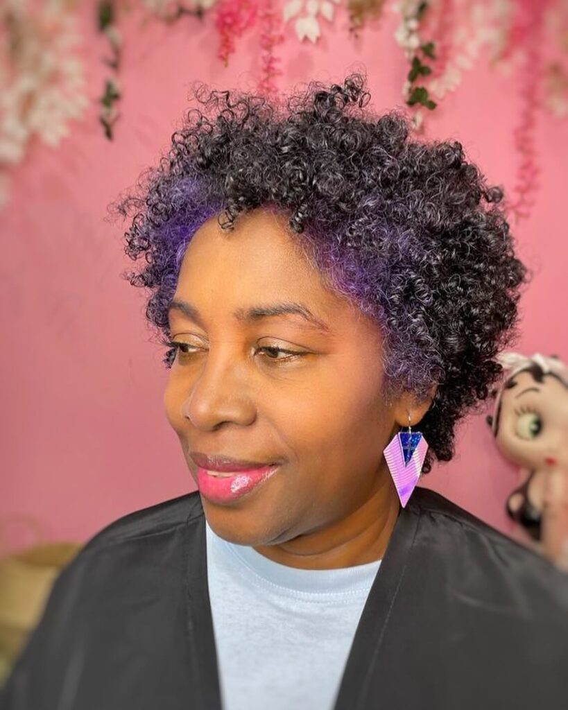 A woman with short tapered dark coils featuring a vibrant purple color pop along the front hairline.