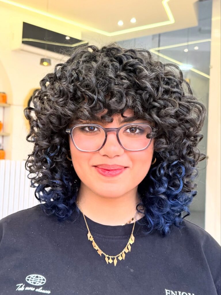 A woman with a voluminous curly shag haircut featuring dark hair with midnight blue dipped ends and curly bangs.