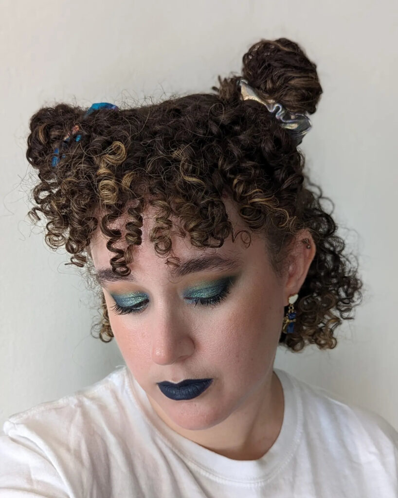A woman with short brown curly hair styled into two half-up space buns with metallic scrunchies and curly bangs.