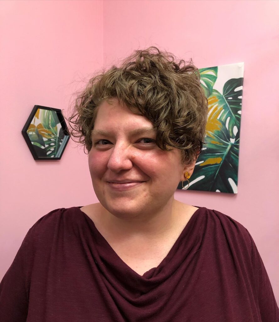 A smiling woman with a messy ash brown curly pixie cut is posing in front of a pink wall.