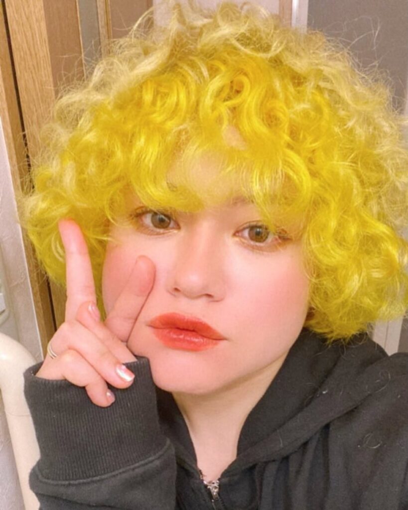 A woman with a chin-length curly bob in a bright neon yellow color and matching curly bangs.
