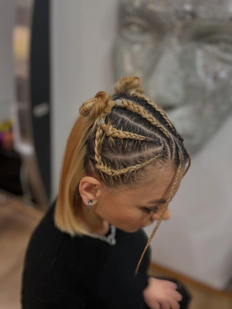A woman with honey-blonde hair featuring intricate, curved scalp braids that lead into two small top buns with silver jewelry accents.