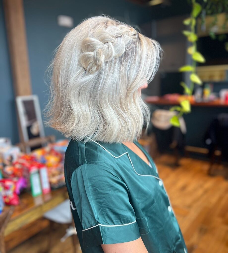 A woman with an icy blonde shoulder-length bob featuring a thick Dutch braid swept to one side across the top of her head.