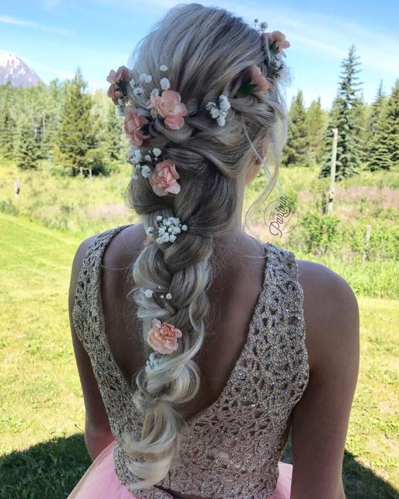 A woman with long ash-blonde hair in a voluminous loose braid adorned with fresh peach flowers and white baby's breath.