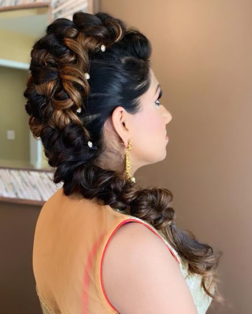 A woman with dark brown and caramel-highlighted hair styled in a voluminous side braid adorned with small sparkling jewels.
