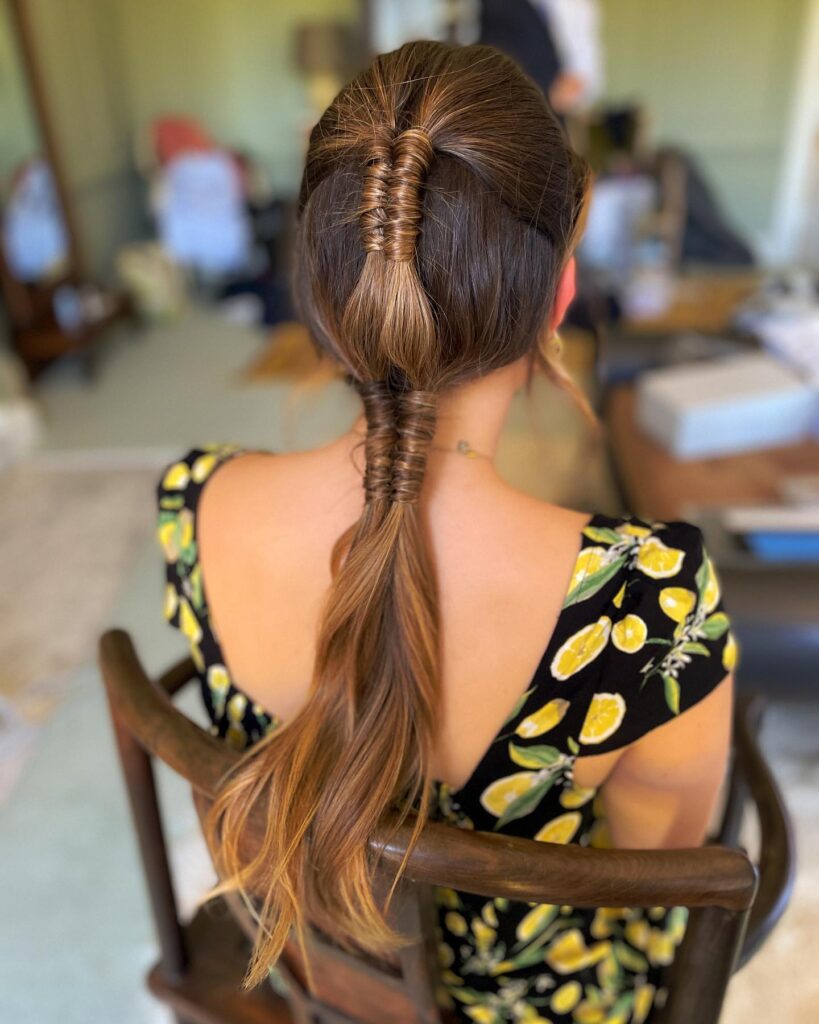 A woman with brunette hair styled in a low ponytail featuring two sections of tight hair-wrapped details along the center.
