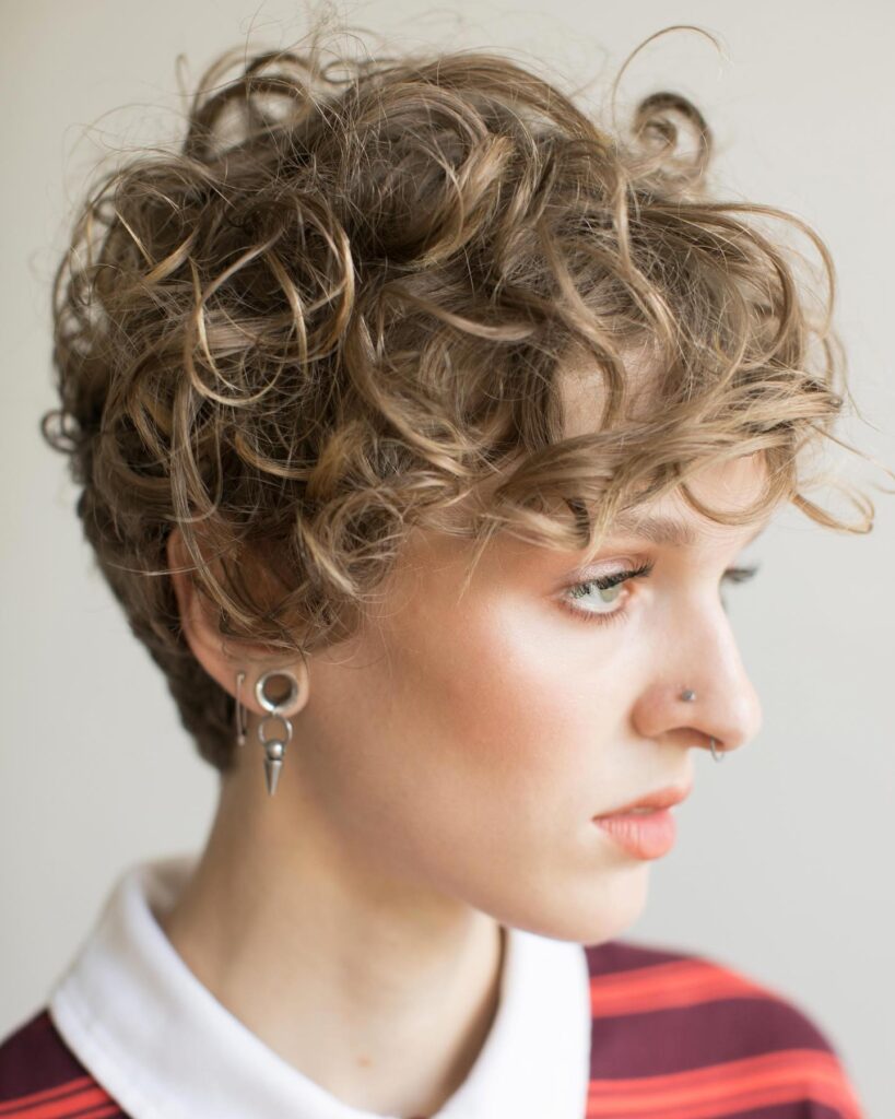 A woman with a dirty blonde curly pixie cut featuring short sides and voluminous ringlets on top that fall forward onto her forehead.