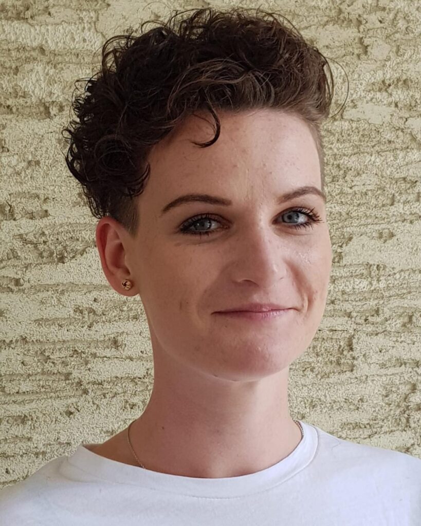 A woman smiling with a dark brown curly pixie cut featuring an undercut on the sides and side-swept voluminous curls on top.