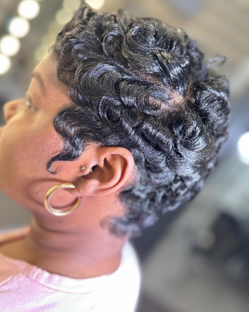 A close-up side view of a woman with a very short jet-black pixie cut styled into deep, glossy finger waves.