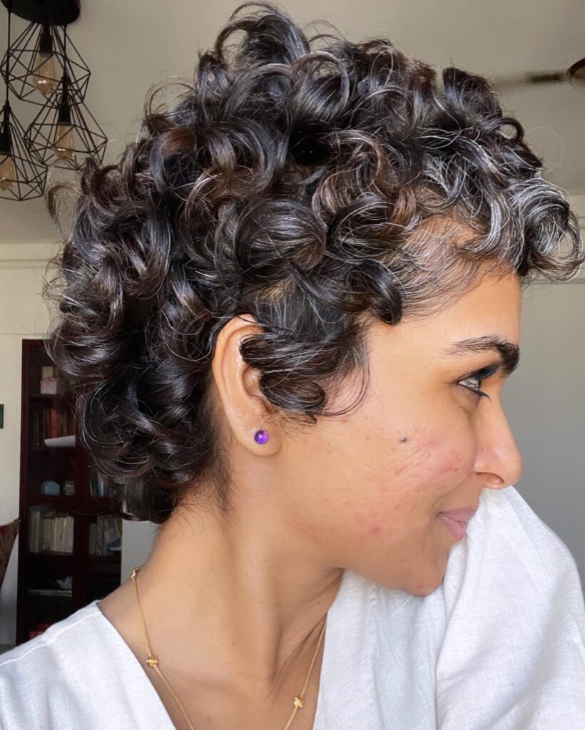 A woman with a salt-and-pepper curly pixie cut featuring tight dark spirals mixed with natural silver hair.