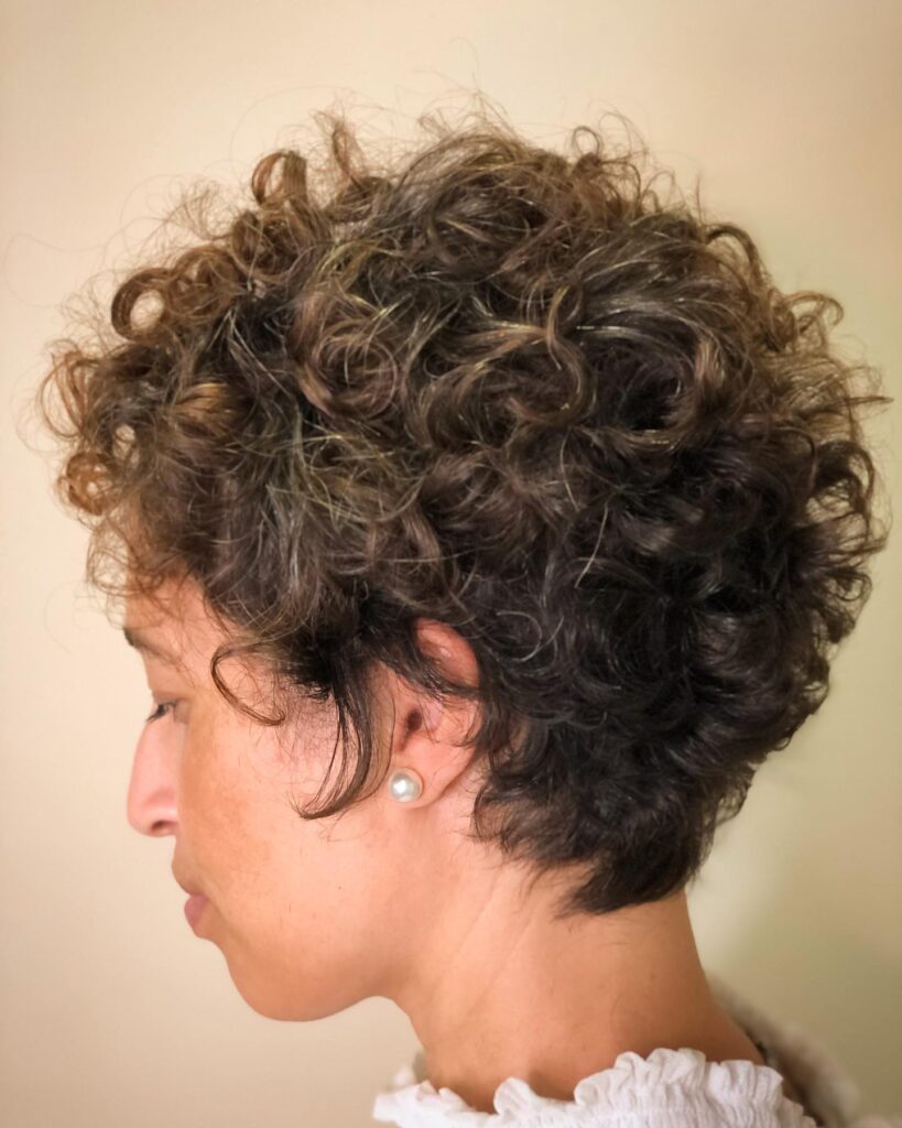 A woman in profile with a messy brunette curly pixie cut featuring soft layers and subtle golden highlights.