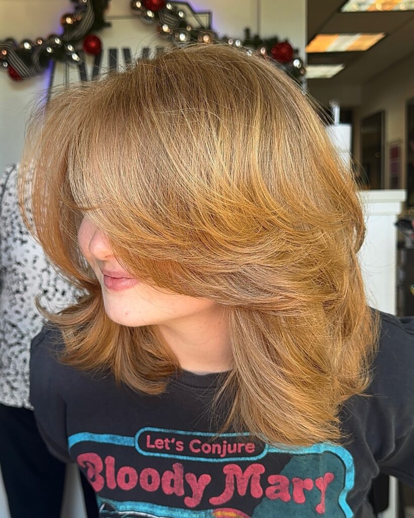 A woman with medium-length honey-blonde hair featuring voluminous feathered layers and long wispy bangs that frame her face.