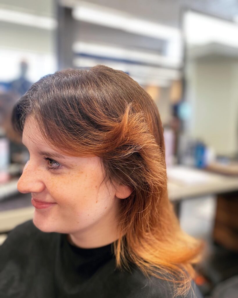 A woman with a dark-to-copper-auburn ombre hairstyle featuring dramatic feathered bangs that sweep outward away from her face.