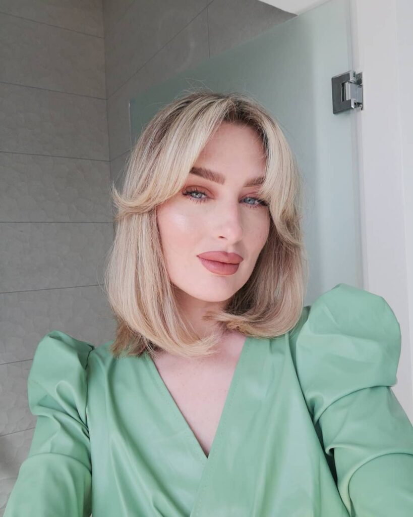 A woman with an ash-blonde shoulder-length bob and long feathered curtain bangs that curve outward to frame her face.