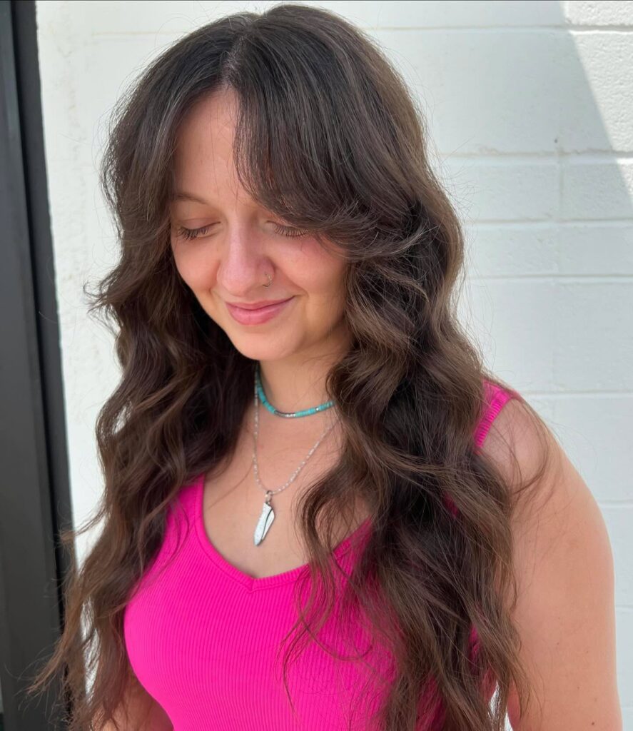 A woman with long, wavy chocolate brown hair and wispy feathered curtain bangs that blend into her face-framing layers.
