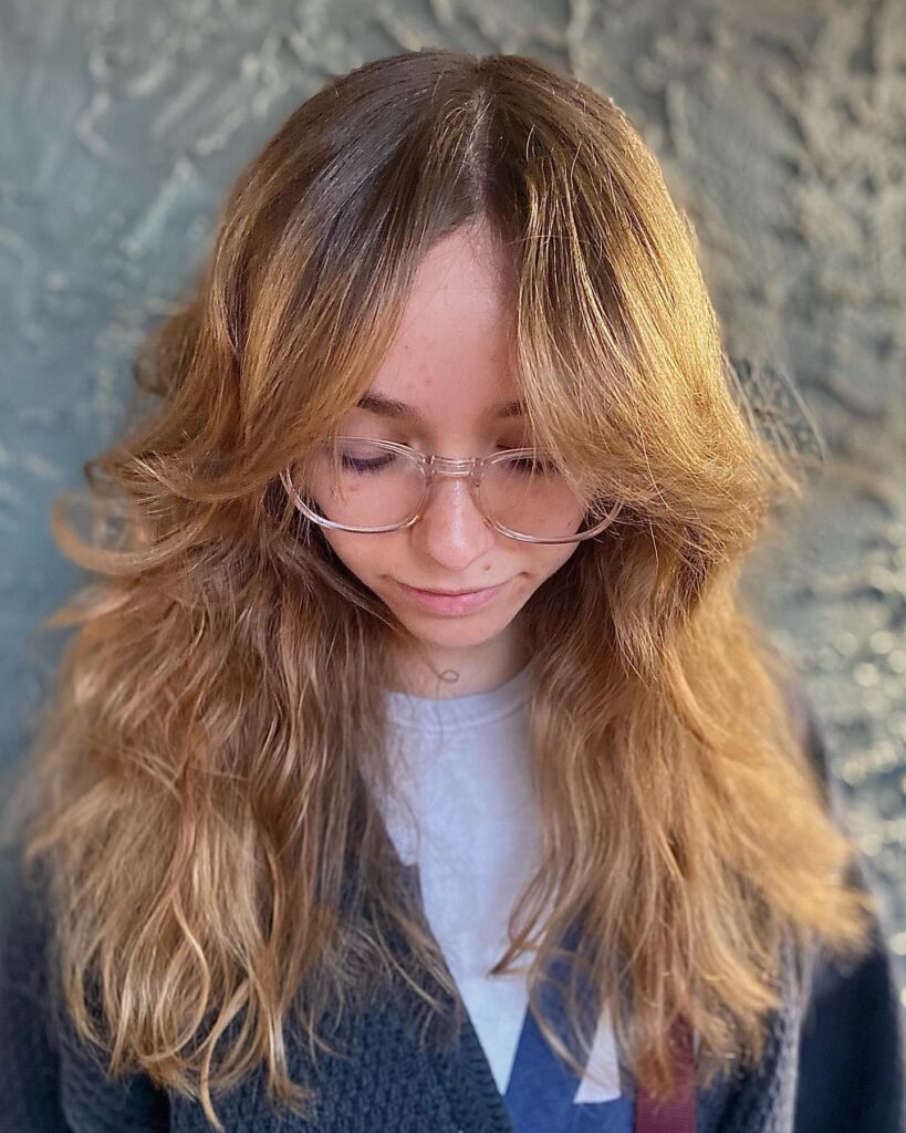 A woman with long, wavy golden-brown hair and feathered curtain bangs that sweep out to frame her face.
