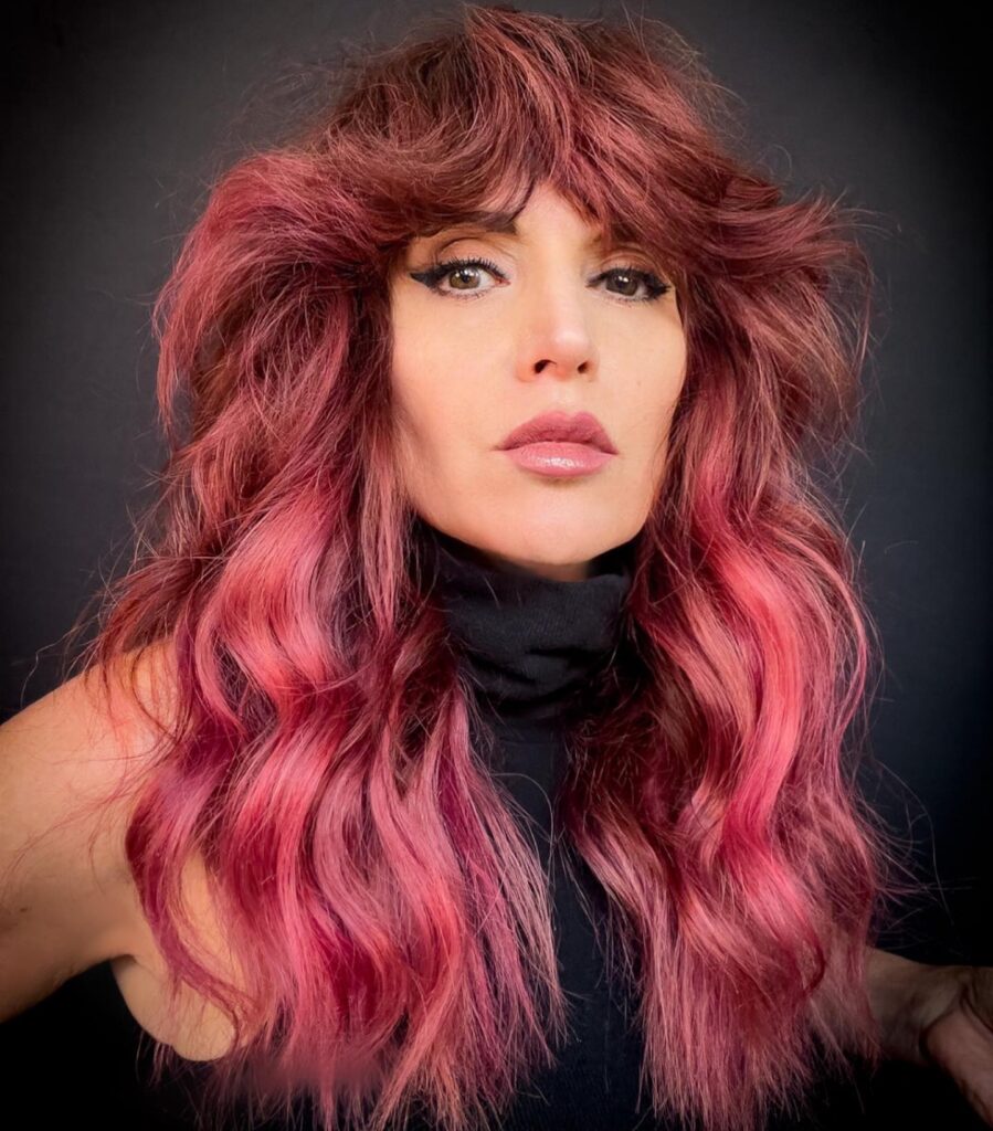 A woman with long magenta and berry-toned hair featuring heavy feathered layers and voluminous bangs that sweep outward.