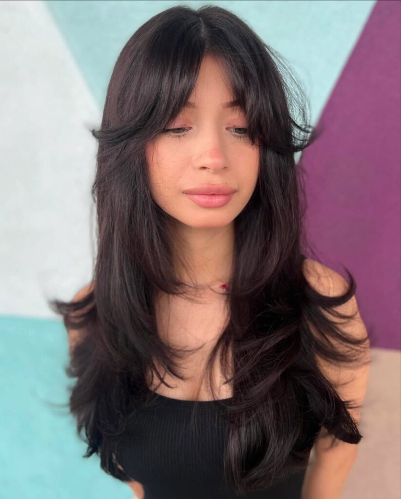 A woman with long, sleek black hair featuring tiered butterfly layers and long feathered bangs that sweep away from her face.