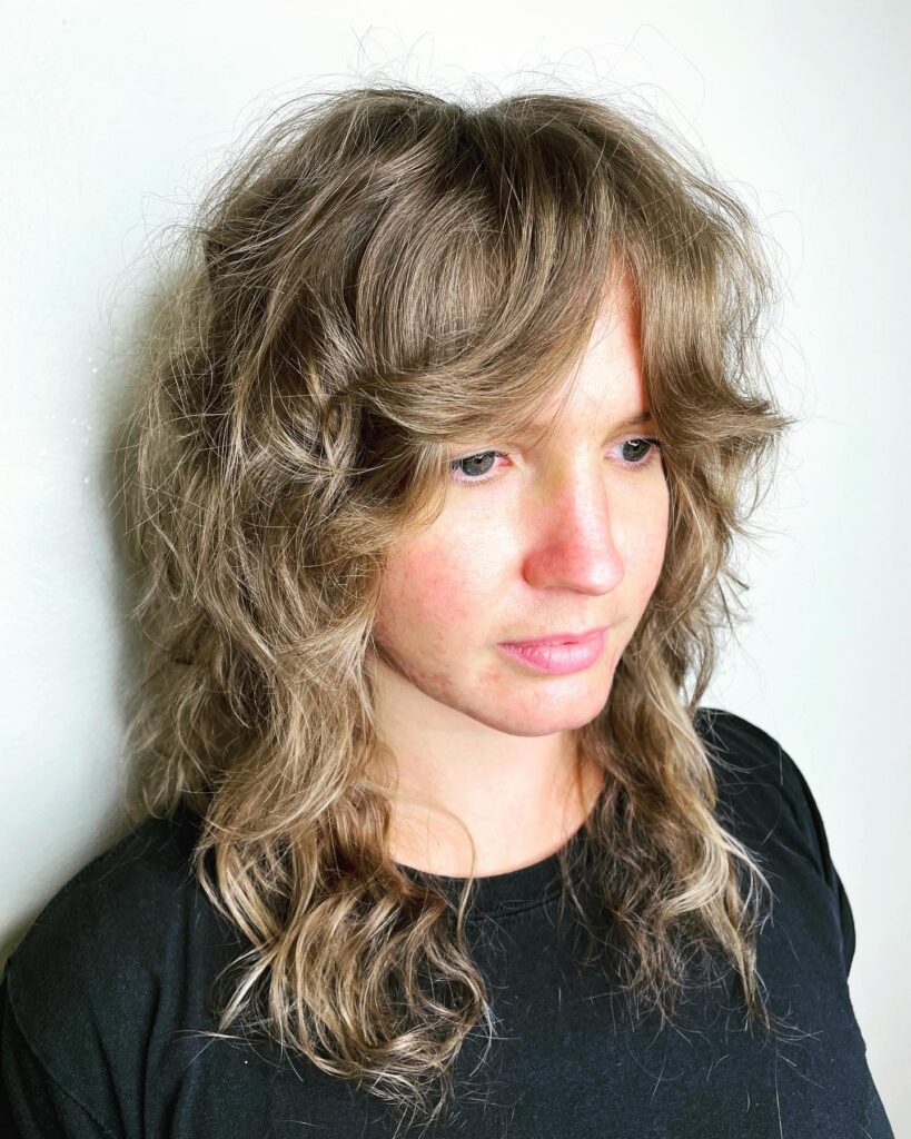 A woman with a messy, ashy brown, shaggy haircut and thick feathered bangs that frame her eyes.