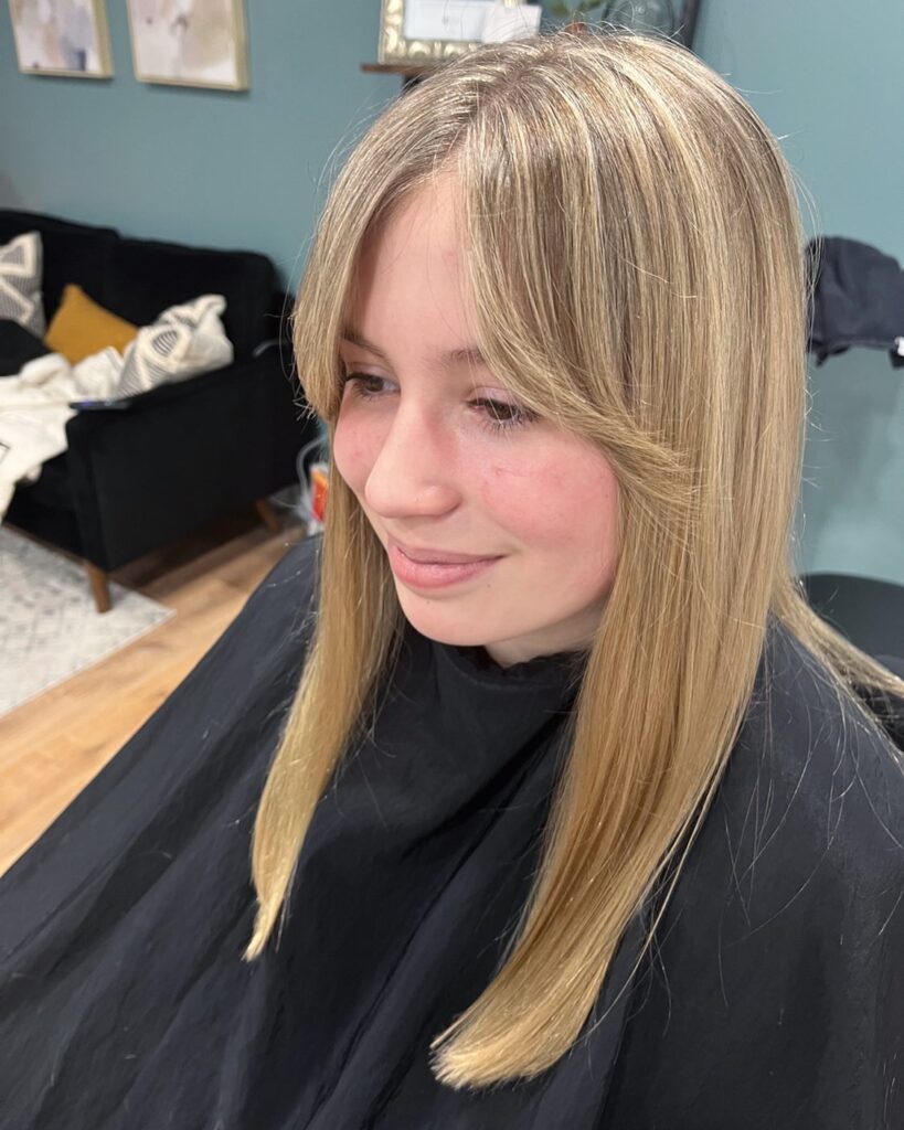 A woman with long, straight sandy blonde hair and soft feathered curtain bangs that blend into her sleek lengths.
