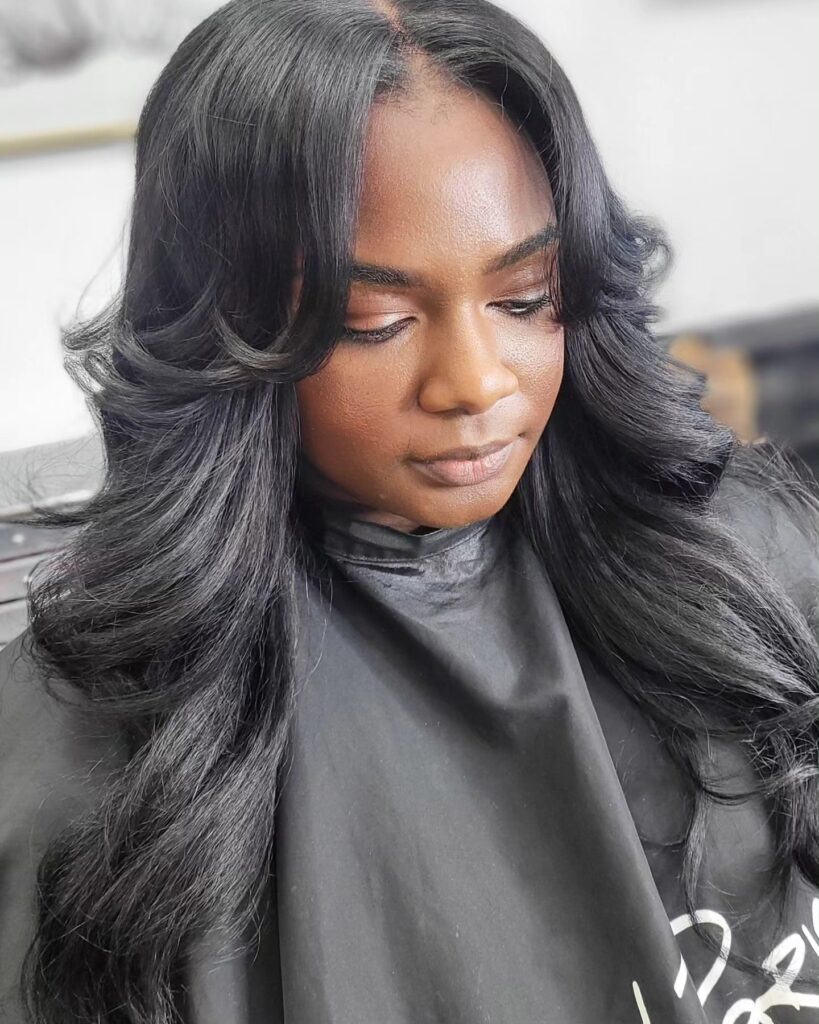 A woman with long, voluminous black hair featuring bouncy curls and perfectly feathered curtain bangs that frame her face.