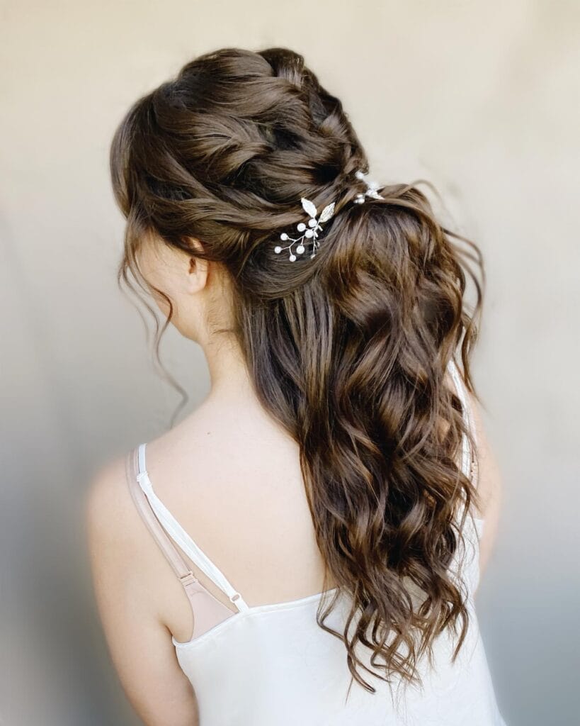 A rear view of a woman showcasing a textured half-up wedding hairstyle on long, wavy brown hair adorned with delicate pearl floral pins.
