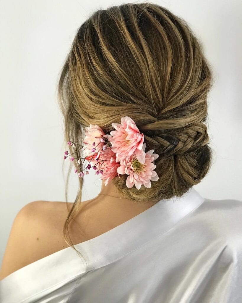A woman with highlighted blonde hair styled in a braided low chignon decorated with fresh pink and purple flowers.
