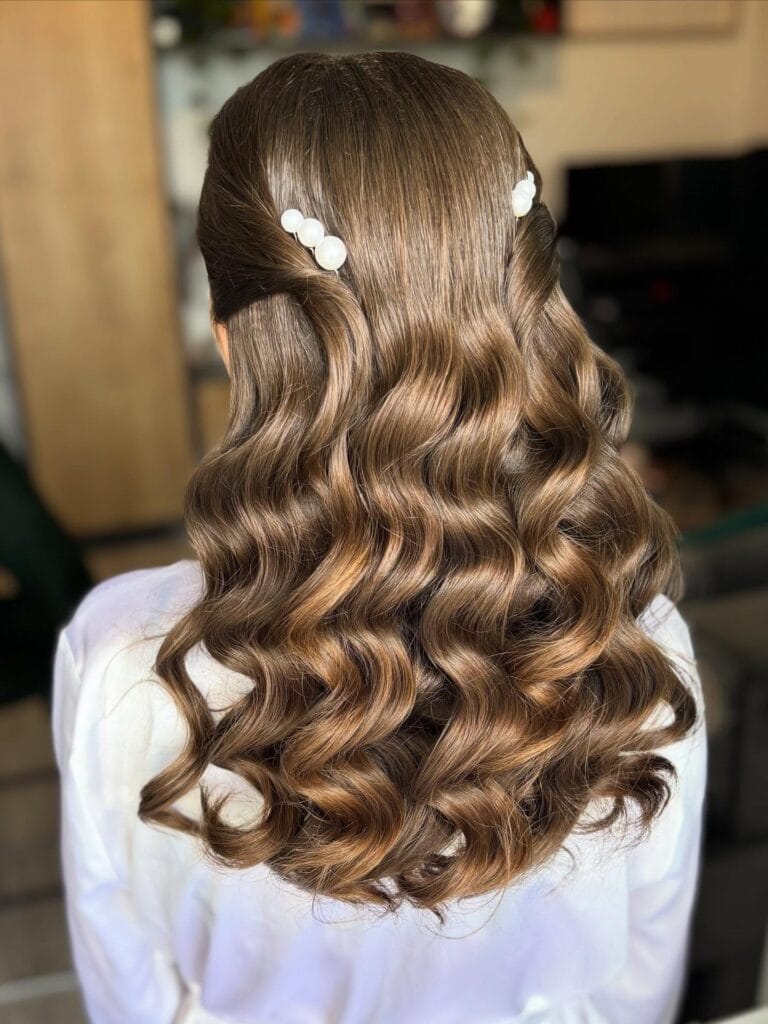 A woman with shiny chestnut brown hair styled in structured S-waves and secured with large pearl clips in a half-up fashion.