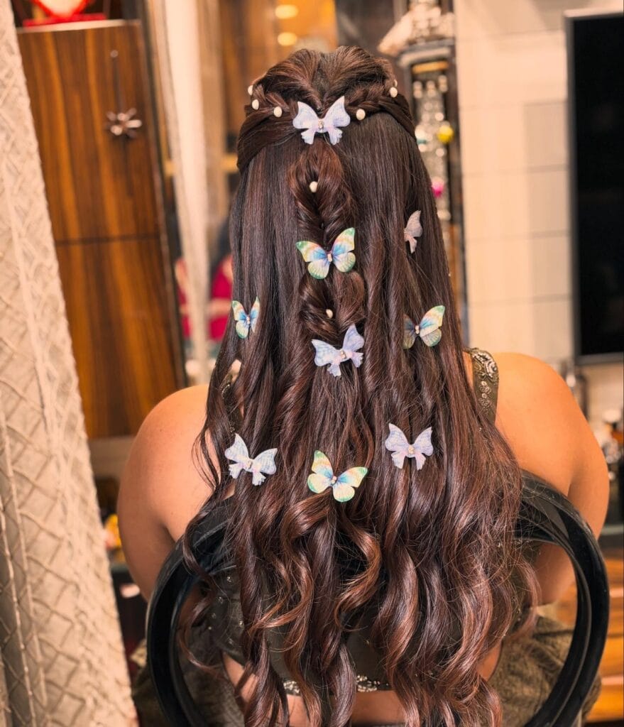 A woman with long wavy chestnut hair featuring a braided half-up hairstyle adorned with colorful butterfly clips and small pearls.