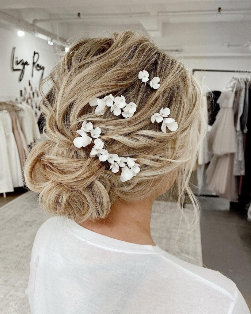 A woman with textured ash-blonde hair styled in a low messy bun decorated with scattered small white 3D flower pins.