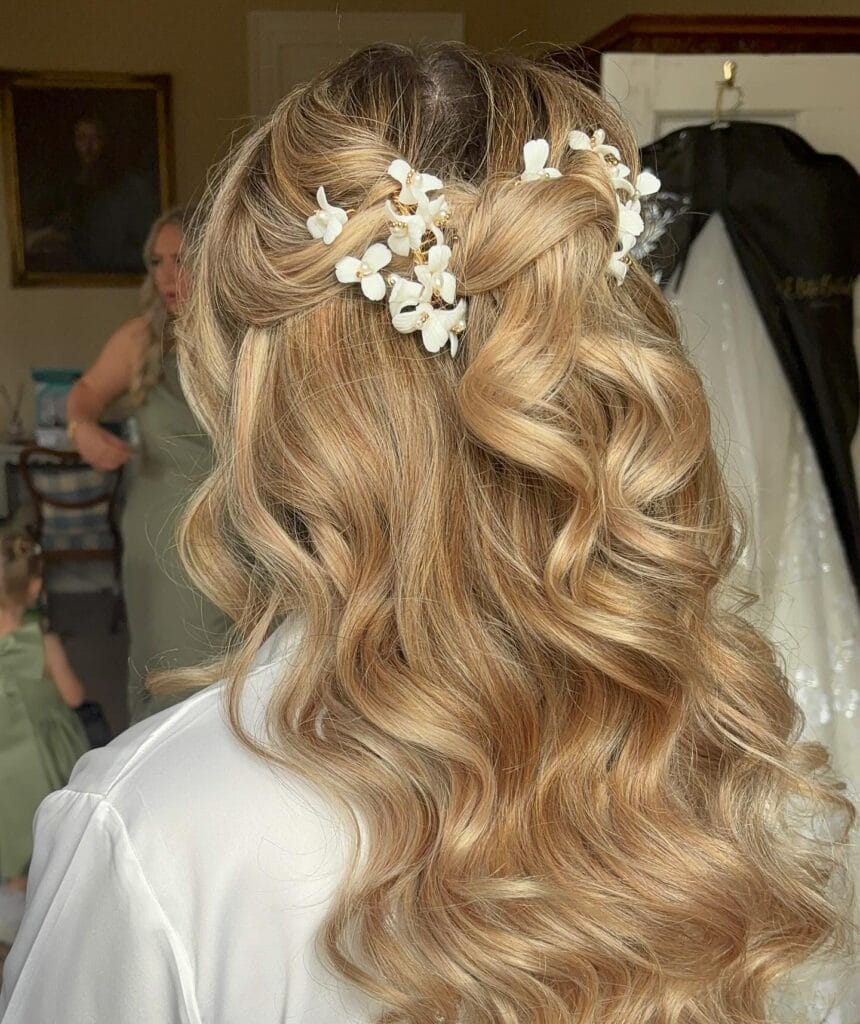 A woman with long, wavy, honey blonde hair featuring a half-up twisted hairstyle adorned with delicate white floral hairpins.