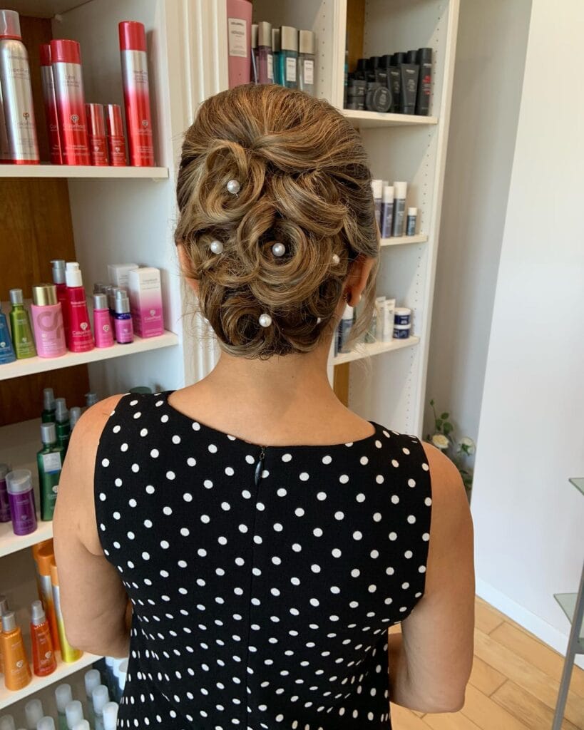 A woman with highlighted brown hair styled into an intricate updo of rose-shaped swirls accented with large white pearls.