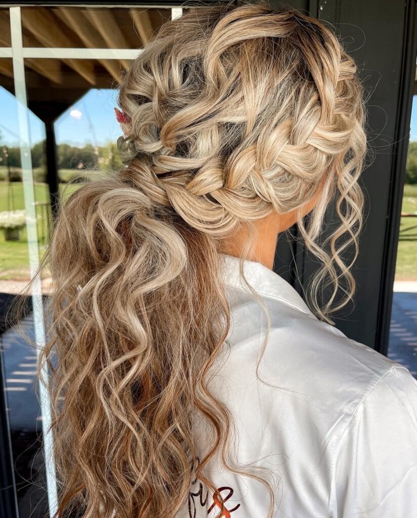 A woman with sandy blonde highlighted hair wears a textured boho hairstyle featuring a side braid tucked into a low curly ponytail.