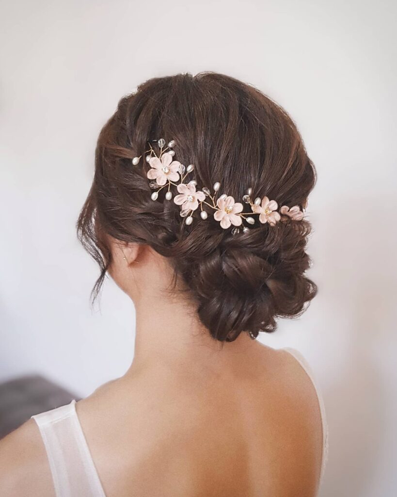 A detailed photo of a woman from behind, showcasing a dark brown, low, twisted chignon hairstyle adorned with a delicate gold wire, pink floral, and pearl hairpiece.