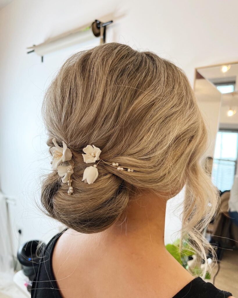 A woman with ash-blonde hair styled in a voluminous low chignon accented with white floral hairpins and loose wavy tendrils.
