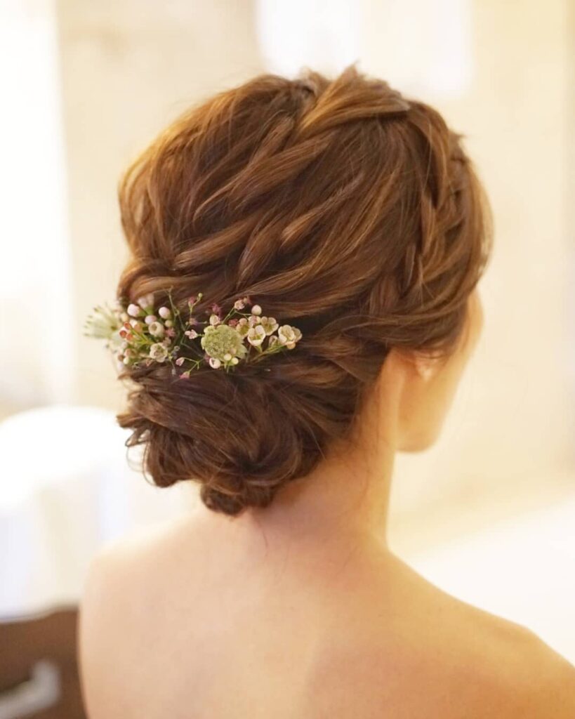 A woman with warm brown hair styled in a braided low chignon decorated with small fresh pink and white wildflowers.