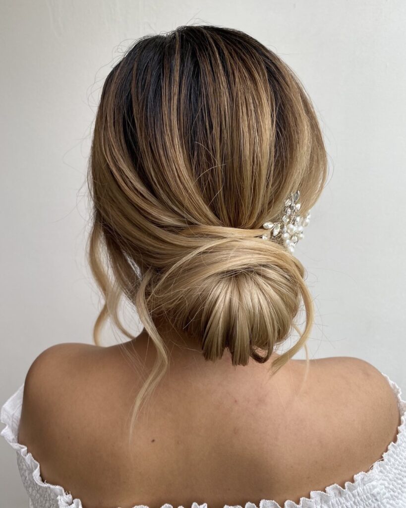 A woman with honey-blonde balayage hair styled into a smooth, wrapped low chignon accented by a pearl and crystal hairpiece.
