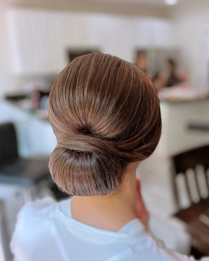 A woman with smooth chestnut brown hair styled in a perfectly polished, minimalist low chignon bun.