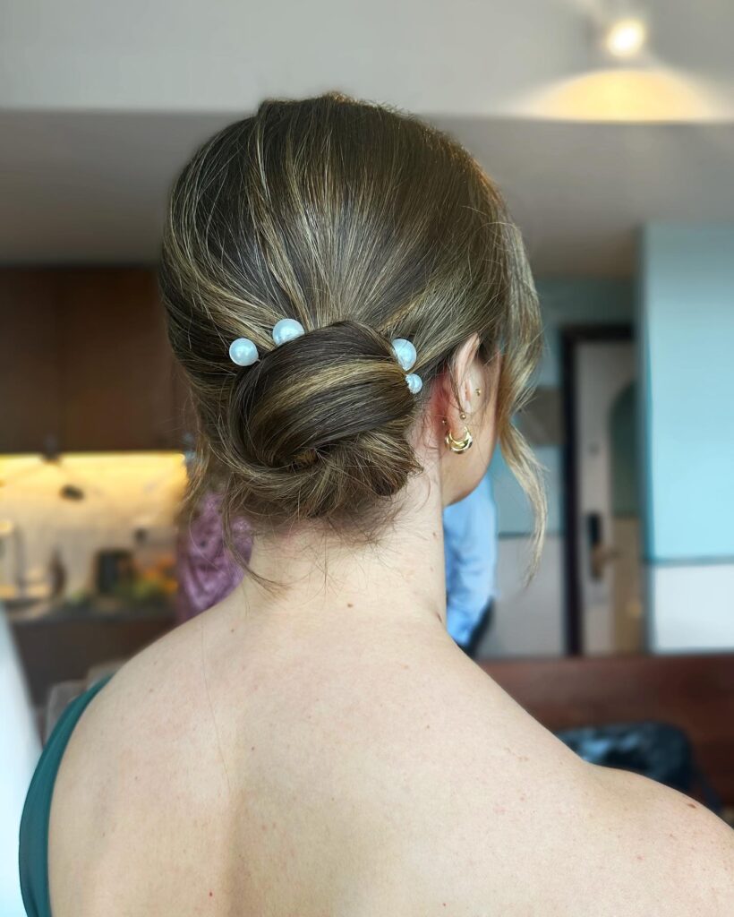 A woman with dirty blonde hair styled in a smooth low chignon accented with several large white statement pearls.