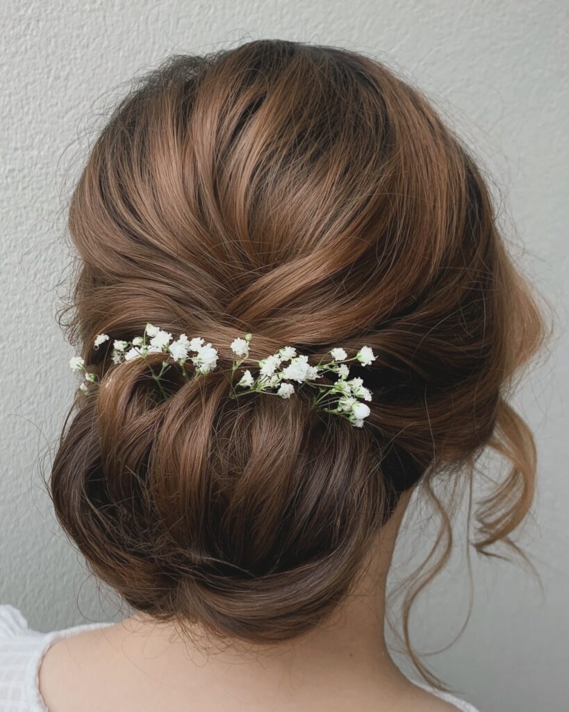 A woman with wavy chestnut brown hair in a loose, low chignon decorated with delicate white baby’s breath flowers.