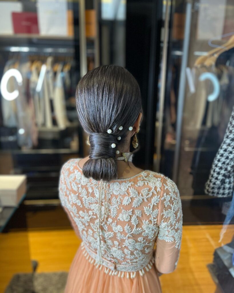 A woman with ultra-smooth dark brunette hair styled in a wrapped low chignon with several white pearls pinned on the side.