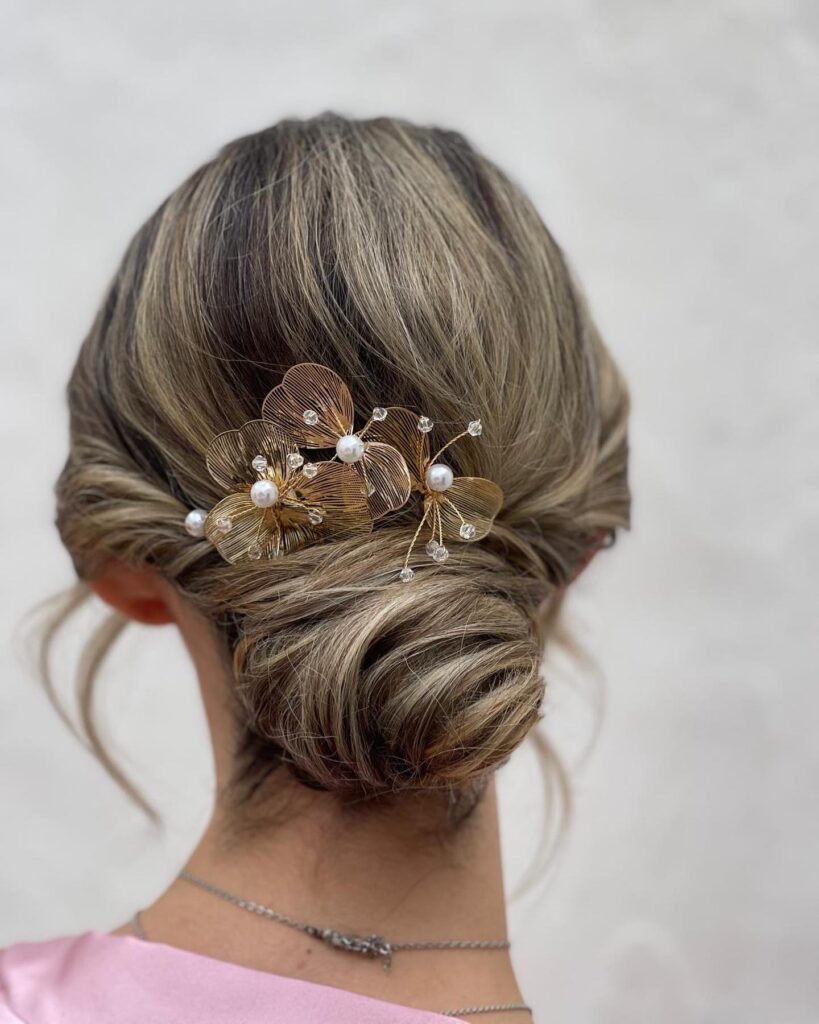 A woman with ash-blonde highlighted hair in a low twisted chignon featuring a large gold wire flower hairpiece with pearls.