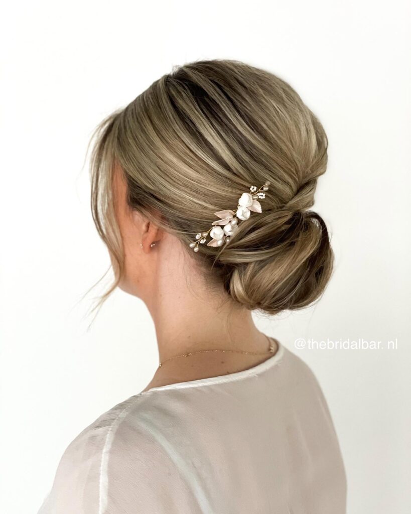 A woman with sandy blonde highlighted hair styled in a smooth tucked low chignon accented by a delicate white flower and pearl hair vine.