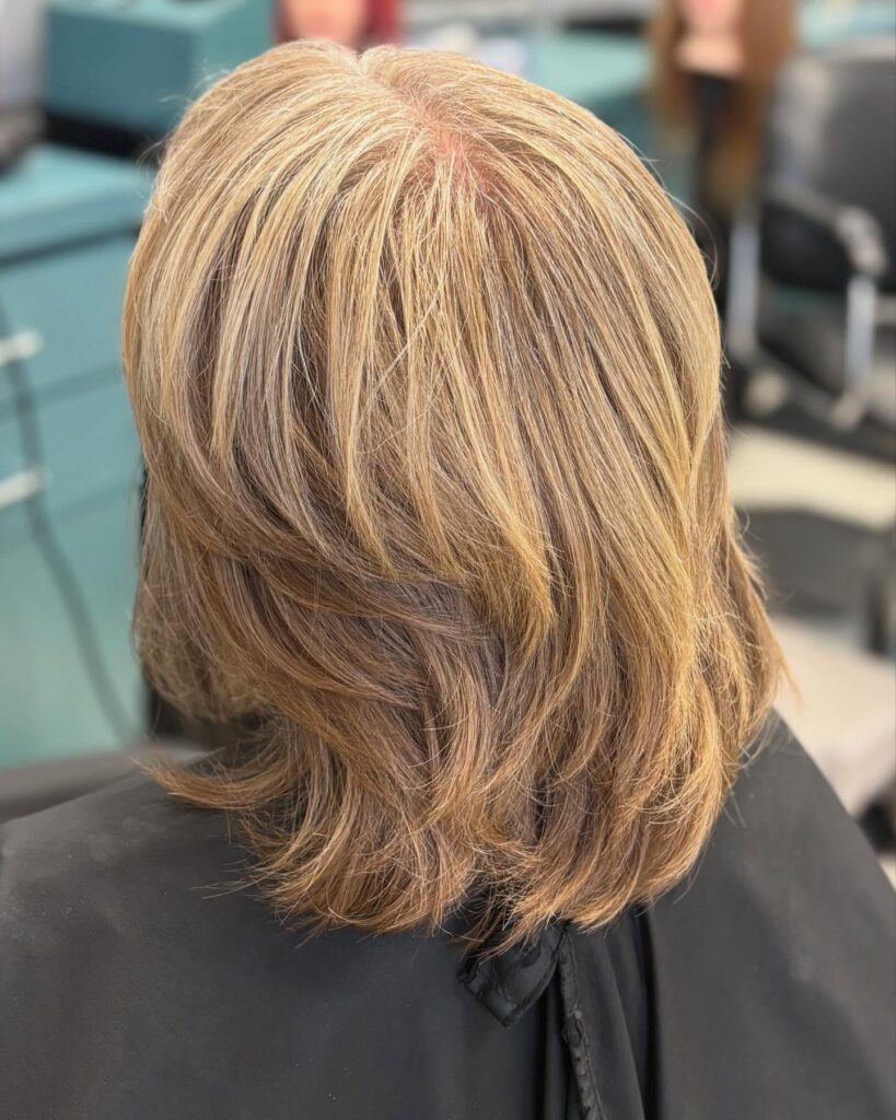A back view of a woman with a shoulder-length honey-blonde lob featuring dimensional highlights and soft, flowing layers.
