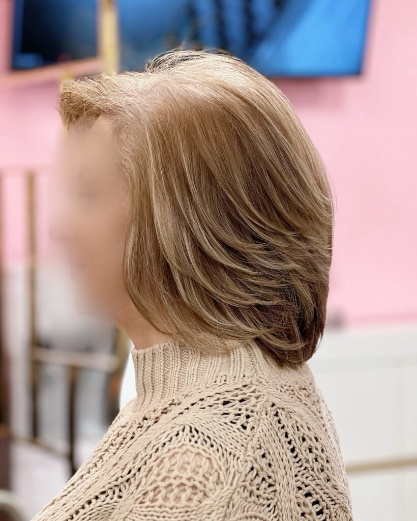 A side profile of a woman with a sandy blonde layered lob featuring bright highlights and a tapered, rounded back.