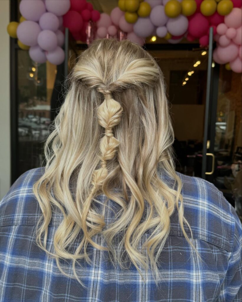 A woman seen from behind with bright blonde highlighted hair in a wavy, medium-length style features a half-up bubble braid down the center.
